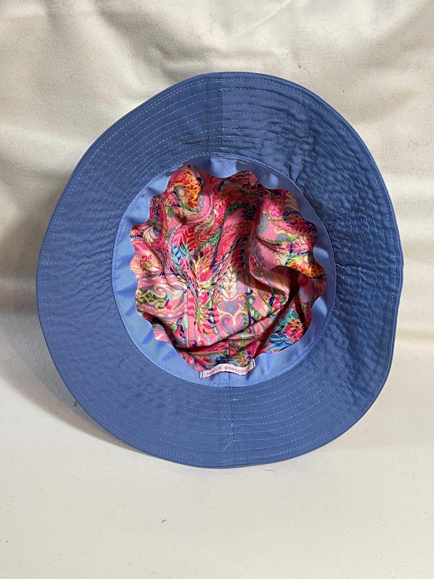 The inside of the bucket hat showing a vibrant pink paisley lining and a tag that reads "HELLO GORGEOUS" sewn to the inside of the brim